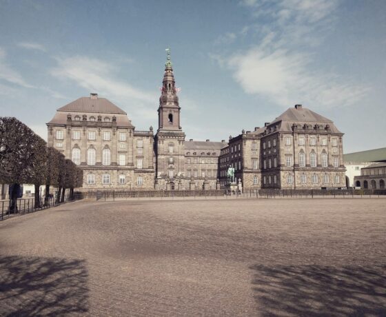 Das Bild zeigt ein imposantes historisches Gebäude mit einem zentralen Turm, das von einer weiten, gepflasterten Fläche und geschnittenen Baumreihen eingerahmt ist. Die klare Perspektive und das sanfte Licht verleihen der Szene eine ruhige, fast majestätische Atmosphäre.