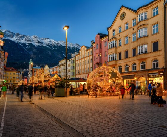 Das Bild zeigt eine weihnachtlich geschmückte Fußgängerzone in Kitzbühel bei Abenddämmerung. Leuchtende Dekorationen, bunte Häuserfassaden und die schneebedeckten Alpen im Hintergrund schaffen eine stimmungsvolle Atmosphäre.
