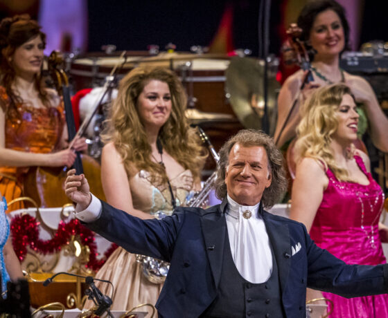 André Rieu beim festlichen Weihnachtskonzert auf der Bühne, umgeben von Musikerinnen in farbenfrohen Abendkleidern. Rieu trägt einen Frack, hält eine Geige in der Hand und breitet lächelnd die Arme aus, während das Orchester im Hintergrund spielt.