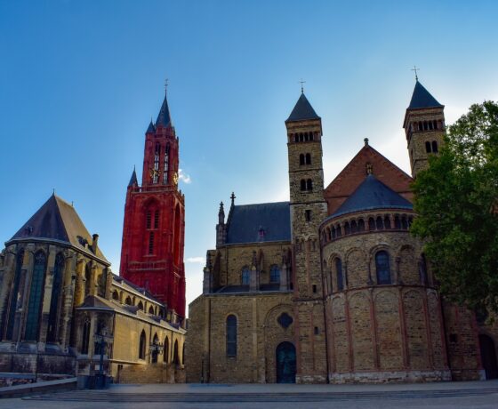 Das Bild zeigt eine eindrucksvolle Kirche mit romanischer und gotischer Architektur. Besonders auffällig ist der rote Glockenturm, der sich markant vom blauen Himmel abhebt und dem historischen Bauensemble eine besondere Note verleiht.