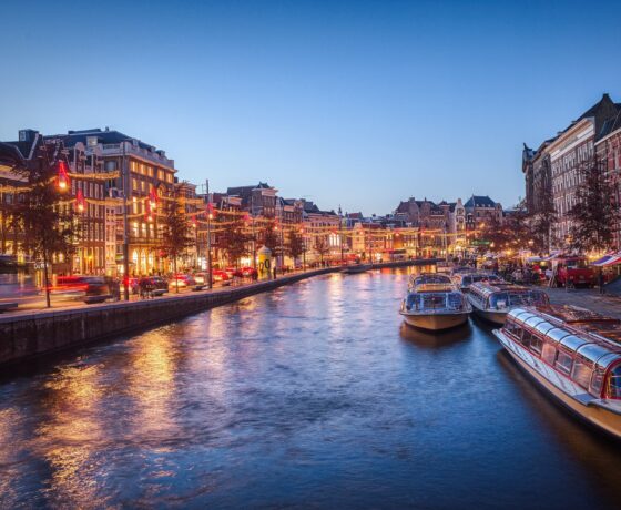 Das Bild zeigt Amsterdam am Abend – beleuchtete Grachtenhäuser, Ausflugsboote und stimmungsvolles Licht entlang des Kanals.
