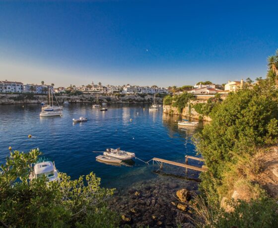 Das Bild zeigt eine ruhige Bucht auf Menorca mit kristallklarem Wasser, kleinen Booten und weißen Häusern im Hintergrund – mediterrane Idylle pur.