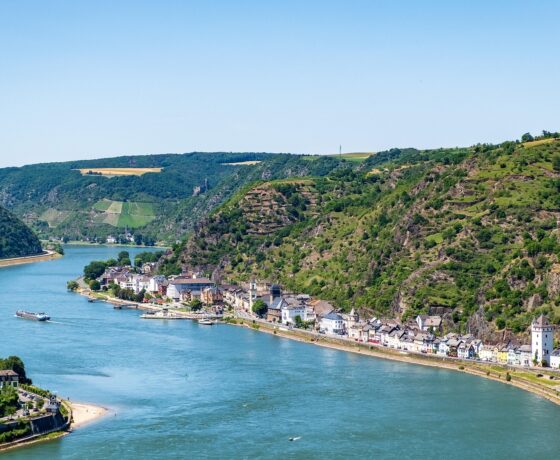 Das Bild zeigt den Rhein, umgeben von steilen Weinbergen und einer historischen Stadt am Ufer mit Fachwerkhäusern und einer Burg auf dem Hügel.