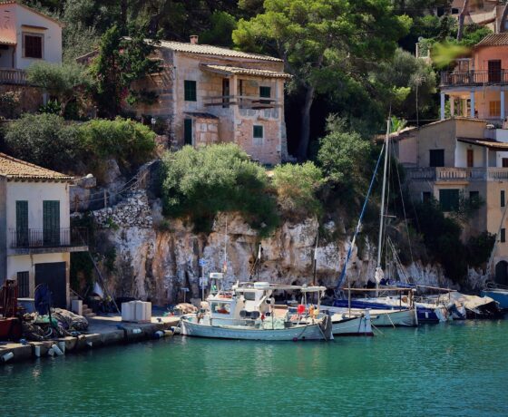 Das Bild zeigt ein idyllisches Fischerdorf auf Mallorca mit Booten im klaren Wasser und charmanten Steinhäusern an felsiger Küste.