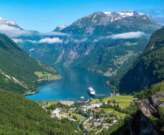 Blick auf den Geirangerfjord mit einem Kreuzfahrtschiff, umgeben von steilen Bergen, grünen Tälern und einem idyllischen Dorf im Vordergrund.