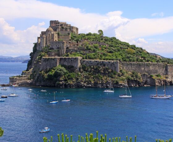 Das Bild zeigt das Castello Aragonese auf Ischia, Italien – eine historische Festung auf einem Felsen im Meer, verbunden mit der Insel durch einen Damm. Boote liegen im klaren Wasser, im Hintergrund sind Berge und Himmel zu sehen.