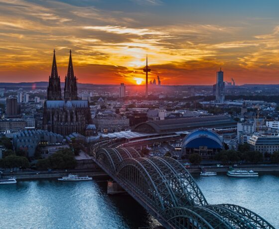 Das Bild zeigt Köln bei Sonnenuntergang mit dem Kölner Dom, der Hohenzollernbrücke und dem Rhein im Vordergrund. Der Himmel leuchtet in warmen Farben.