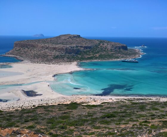 Das Bild zeigt die Küste Kretas mit klarem, türkisfarbenem Wasser, weißem Sandstrand und einer felsigen Halbinsel. Im Hintergrund ist das offene blaue Meer zu sehen.
