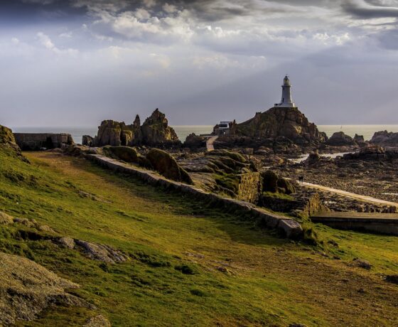 Grüne Küste mit Felsen und weißem Leuchtturm auf Jersey bei bewölktem Himmel.