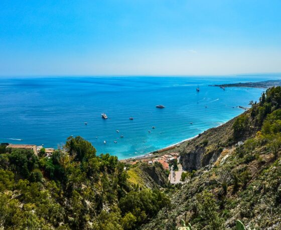 Das Bild zeigt die Küste von **Sizilien** mit blauem Meer, kleinen Booten und grünen Hügeln im Vordergrund.