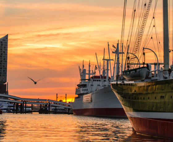 Hamburger Hafen bei Sonnenuntergang mit der Elbphilharmonie im Hintergrund und historischen Schiffen im Vordergrund.