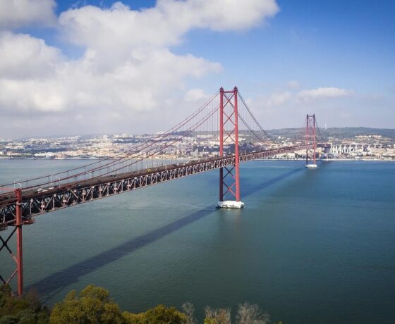 Das Bild zeigt eine große rote Hängebrücke, die sich über einen breiten Fluss erstreckt, mit einer Stadt im Hintergrund unter blauem Himmel mit einigen Wolken.
