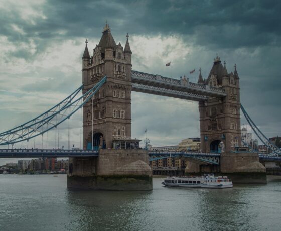 Das Bild zeigt die Tower Bridge in London, eine ikonische Klappbrücke über die Themse, bei bewölktem Himmel. Ein Boot fährt unter der Brücke hindurch.