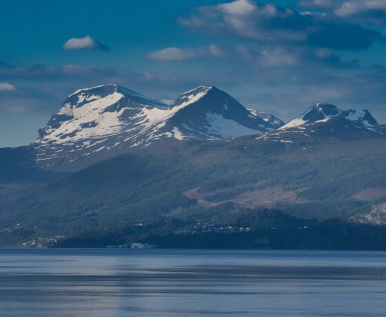 Das Bild zeigt schneebedeckte Berge über einem ruhigen Fjord oder See, mit teils bewaldeten Hängen unter blauem Himmel mit vereinzelten Wolken.
