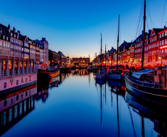 Abendstimmung im Hafenviertel mit beleuchteten Häusern, festgemachten Segelbooten und ruhigem Wasser, das die Lichter stimmungsvoll spiegelt.