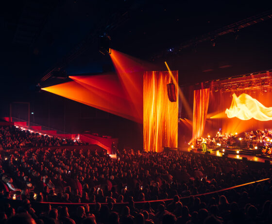 Konzertsaal mit großem Publikum, dramatischer Lichtshow in Orange- und Rottönen sowie einer Live-Bühnenperformance mit Musikern und Sänger*innen.