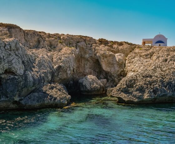 Das Bild zeigt eine felsige Küste mit kristallklarem, türkisfarbenem Wasser. Am rechten Bildrand ist ein weiß-blaues Haus zu sehen, typisch für mediterrane Architektur.