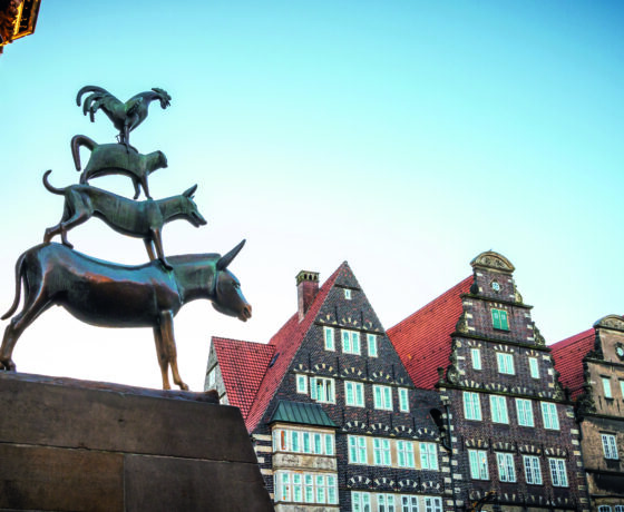 Das Bild zeigt die Bremer Stadtmusikanten vor dem strahlend blauen Himmel und historischen Fachwerkhäusern.